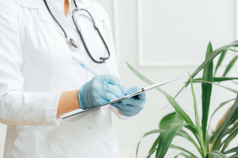 A clinician wearing a white lab coat and stethoscope stands holding a clipboard in gloved hands next to an indoor plant