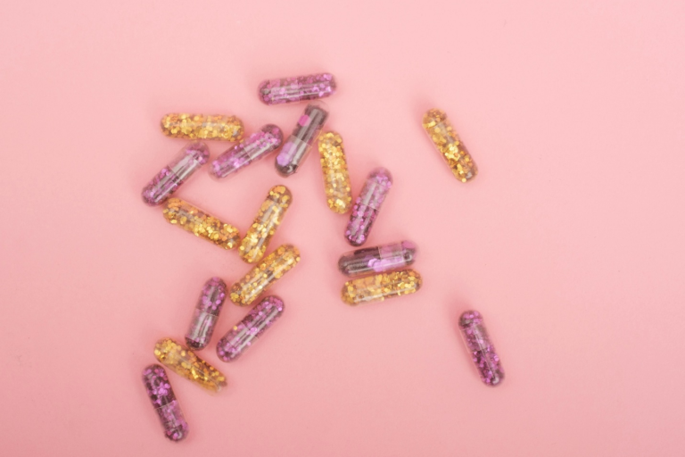 Capsules with gold and lilac glitter sit on a light pink surface