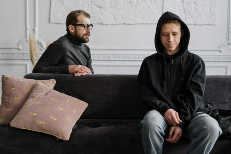 A teenage child in a black hoodie and jeans sits on a grey sofa with pink cushions while an adult in a grey polo neck and spectacles sits behind him against a white textured wall with moldings