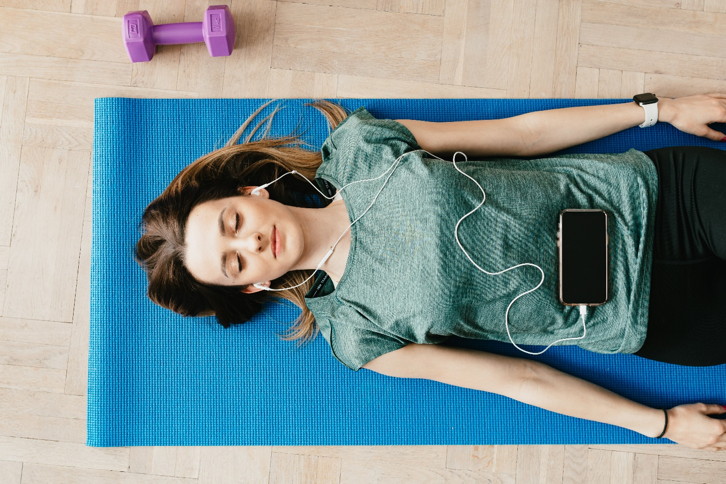 A woman lies on a blue yoga mat beside a dumbbell with earphones in. Her phone lies on her stomach, and she explores the benefits of mindfulness in Miami’s addiction therapy.