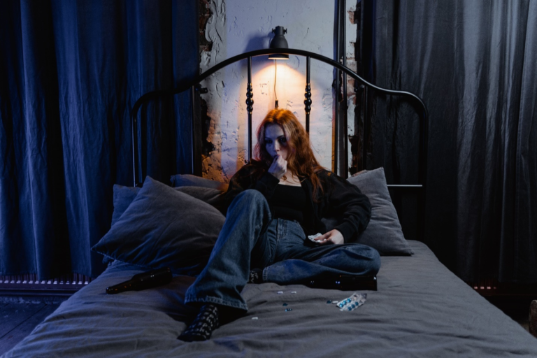 A girl in distress sits on a dimly lit bed surrounded by pills and an empty beer bottle, highlighting the struggles of substance abuse and the need for intervention leading to long-term recovery.