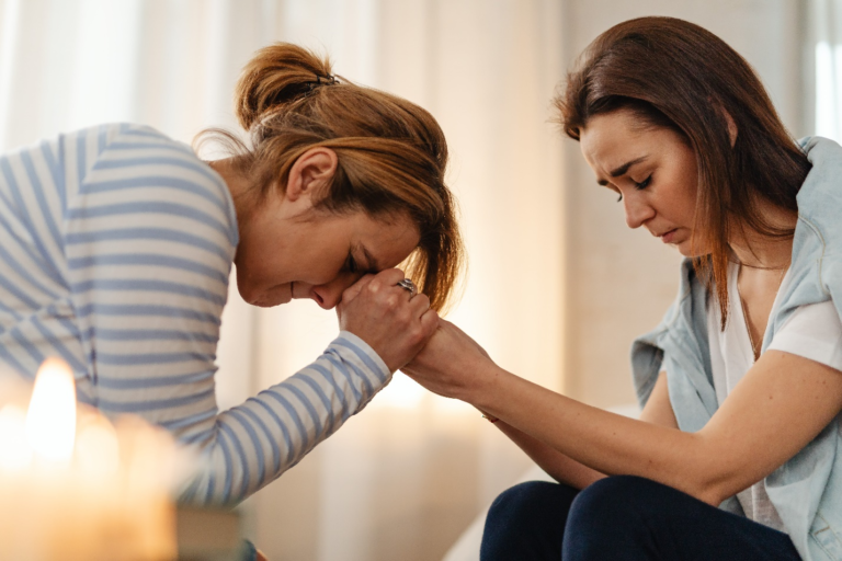 A woman in a blue and white striped t-shirt holds the hands of another woman in a blue shirt while facing down with her lips pursed, showing the support needed to overcome opioid dependency.