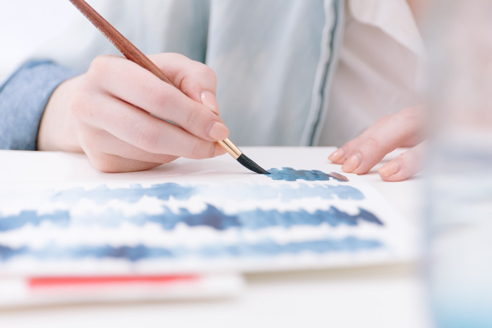 A close-up of a person's hand holding a paintbrush, creating blue watercolor strokes on paper.