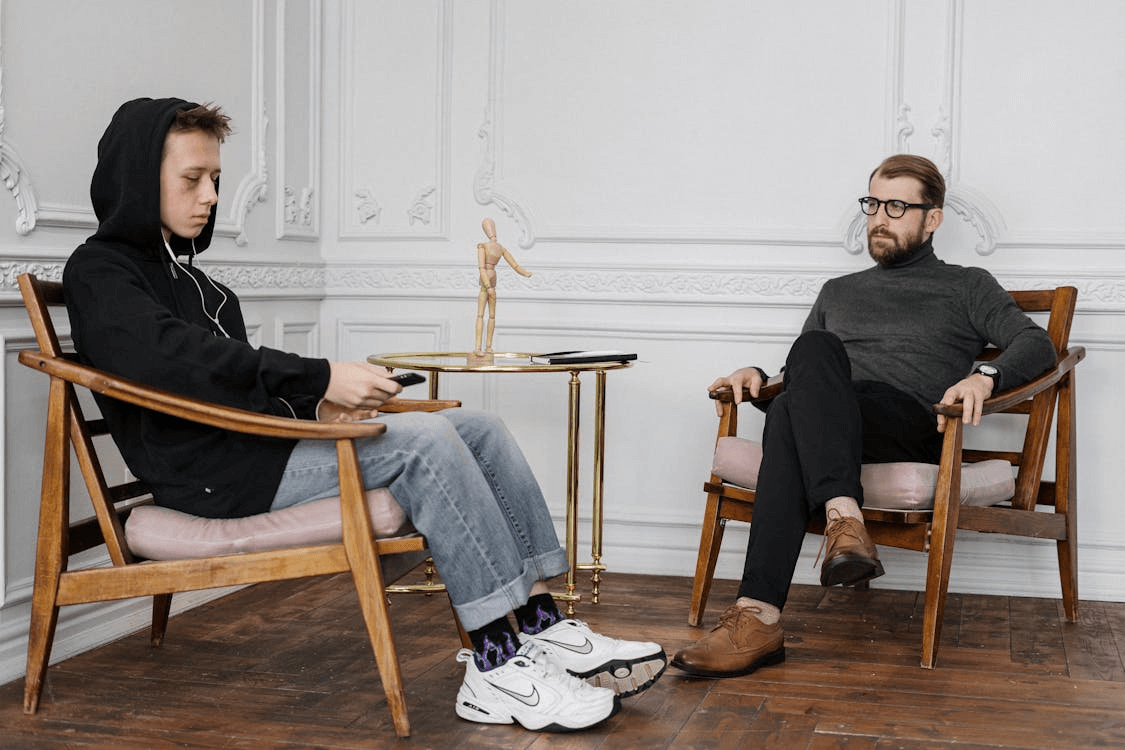 A young person in casual attire sits on a wooden chair, while an older man in formal clothing observes him in a modern, minimalist room.