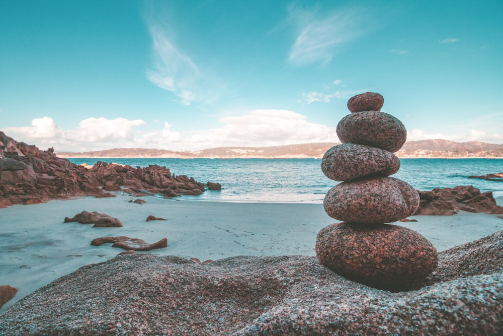 A tall cairn of six round stones balanced on a rocky beach with the ocean and a blue sky in the background.