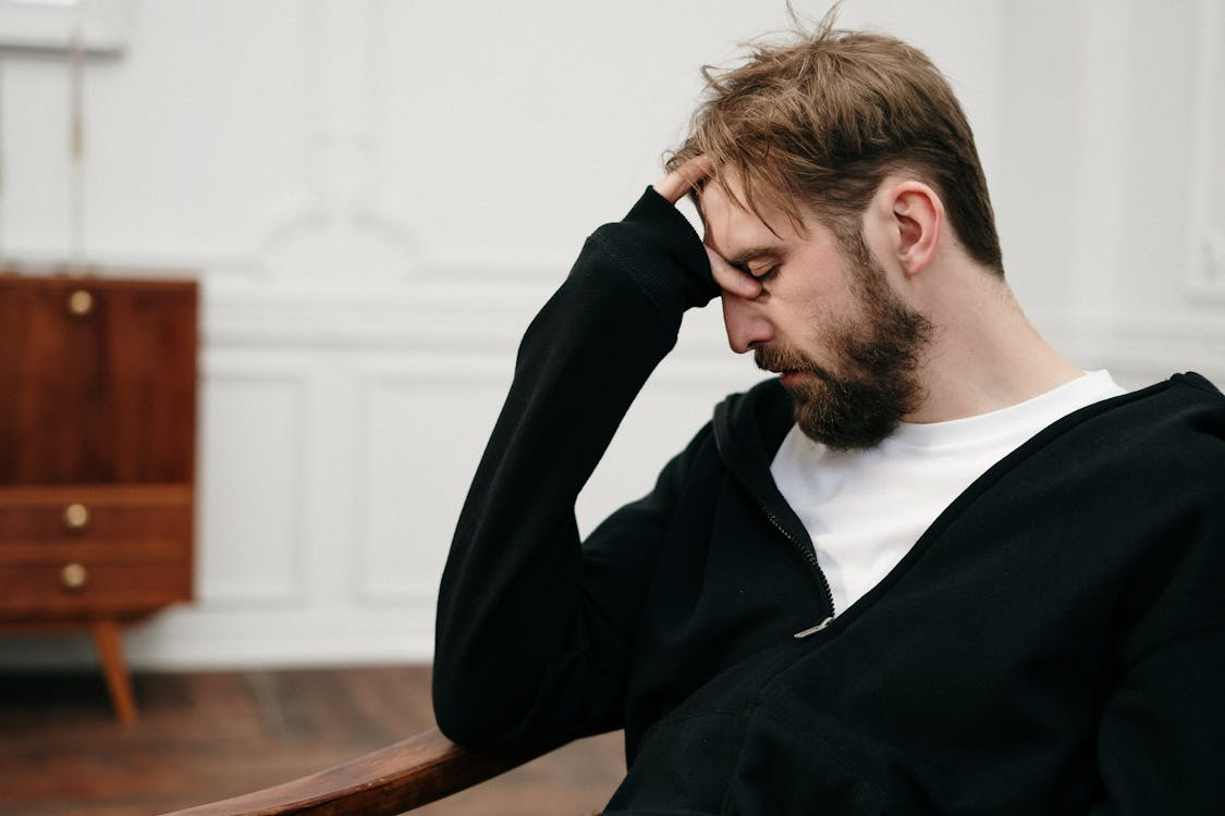 A man in a black hoodie is sitting with his hand on his forehead, appearing deep in thought or stressed.