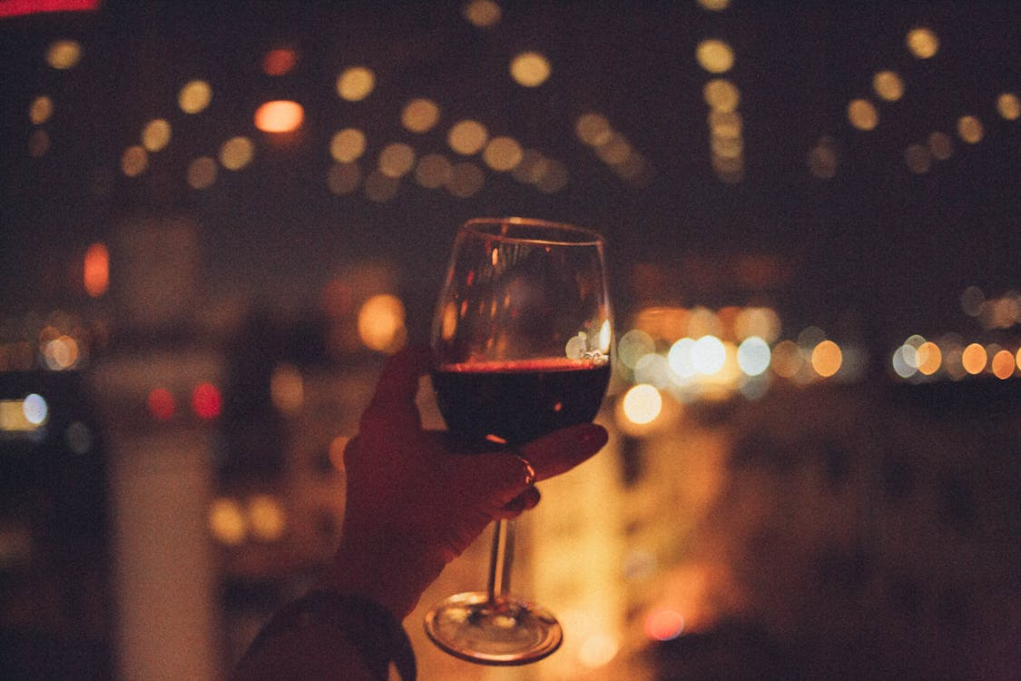 A person holding a glass of red wine with a blurred city background at night.
