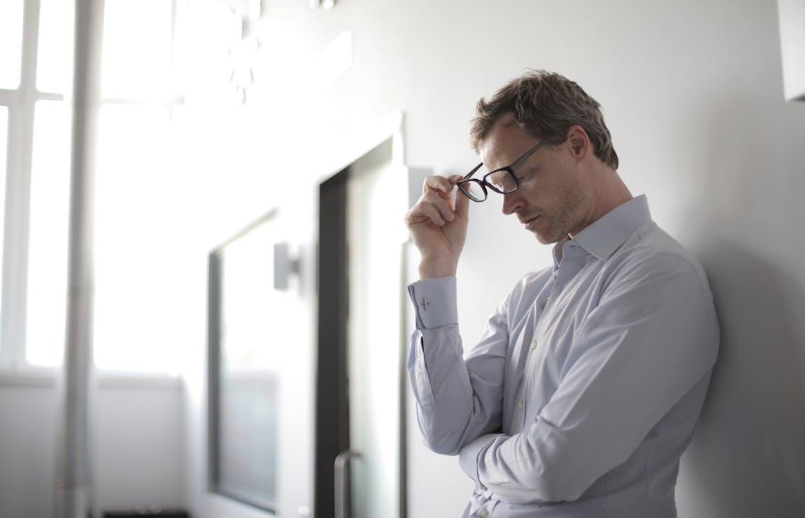 A man standing with his hand on his glasses, appearing deep in thought in a bright, modern room.