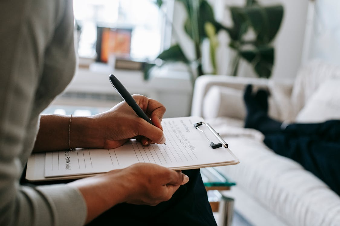 A therapist writes notes on a clipboard during a session with a client relaxing on a couch.