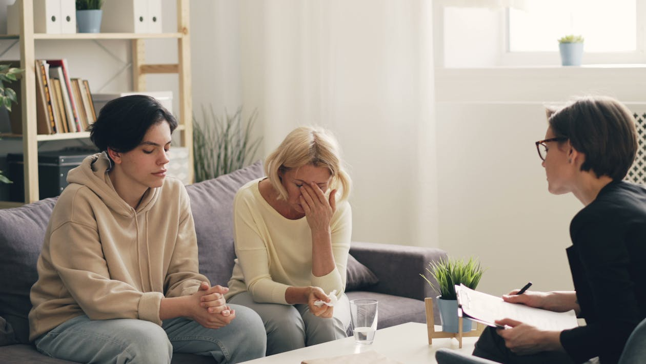 A woman is wiping her face with a tissue while sitting alongside another woman, during a therapy or counseling session with a therapist.