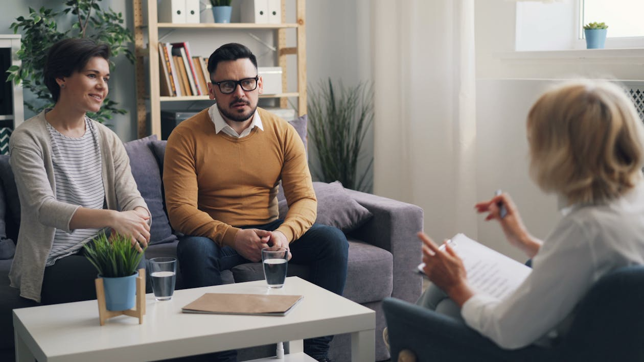 A couple sitting together on a sofa during a therapy session with a counselor, who is taking notes.