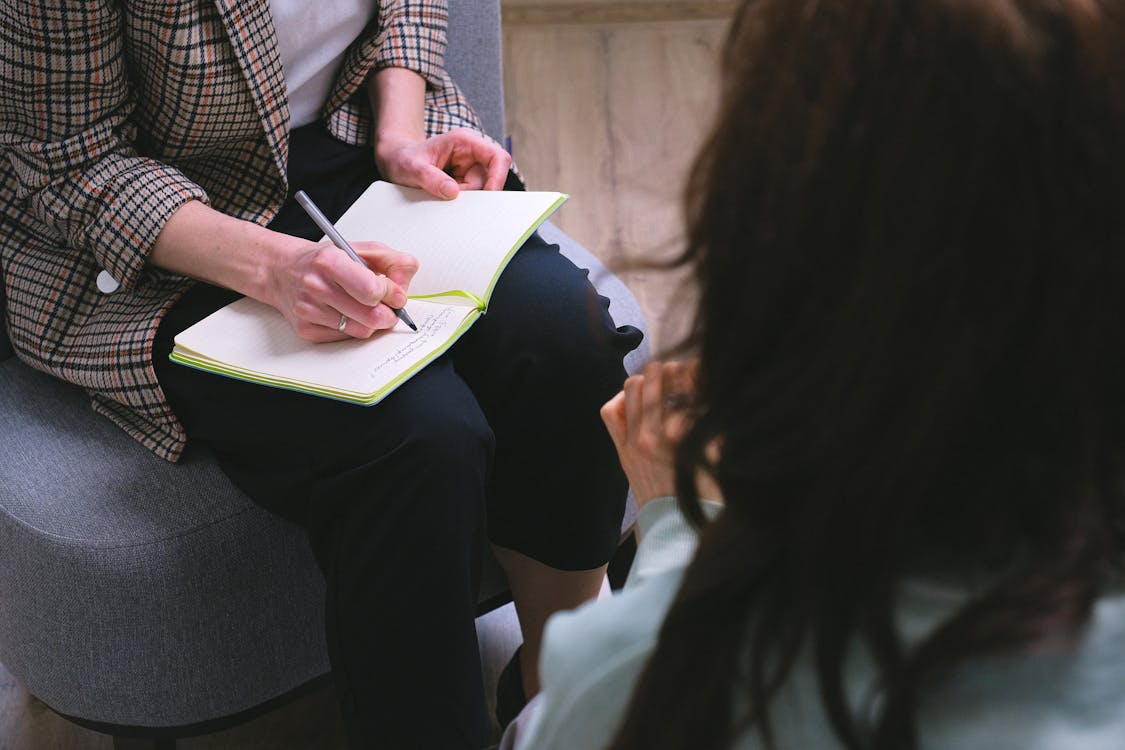 A therapist writes notes in a notebook while listening to a client during a counseling session.
