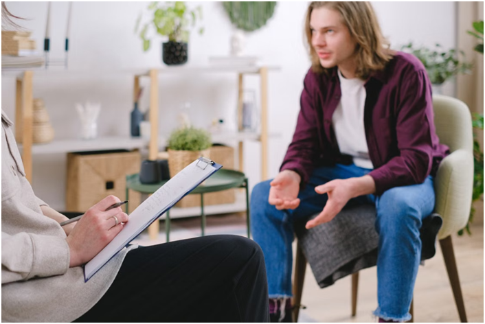 Young man with long hair gesturing while speaking to a therapist writing notes on a clipboard